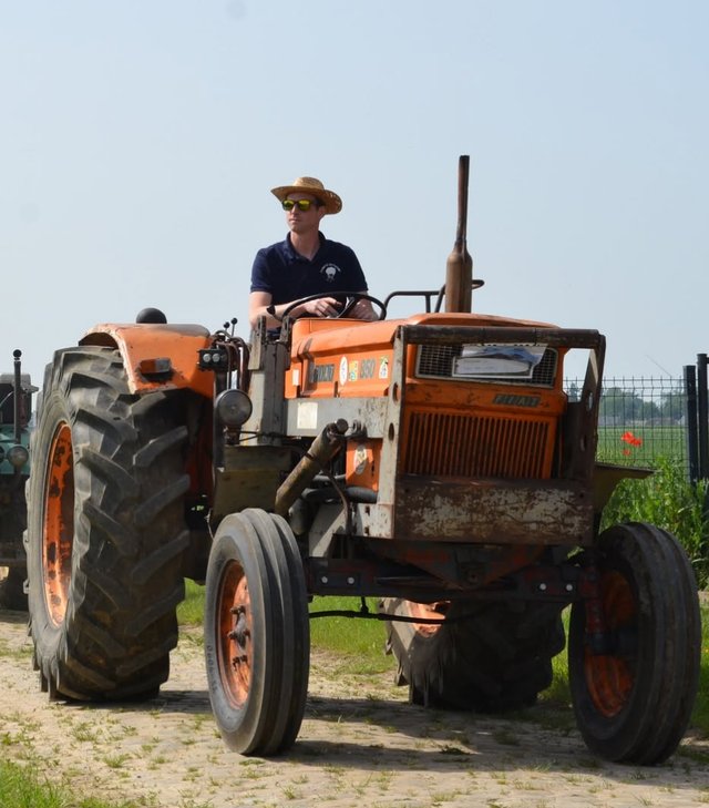 Geoffrey Brassinne au volant d'un vieux tracteur orange, chapeau de paille et lunettes de soleil, sous le soleil d'été.